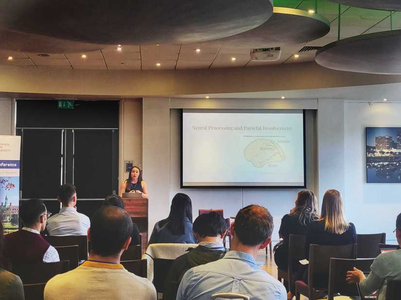 A person is standing at a podium; in the background there is a large screen showing an image; the audience is sitting facing the person presenting.